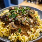 A close-up of a creamy beef stroganoff skillet meal served over egg noodles and garnished with fresh parsley.