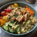 A vibrant and healthy salmon power bowl featuring quinoa, cucumber, tomatoes, shredded carrots, and a creamy dressing.