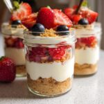 Close-up of High Protein Cheesecake Jars layered with graham cracker crumbs, creamy filling, and fresh strawberries and blueberries.
