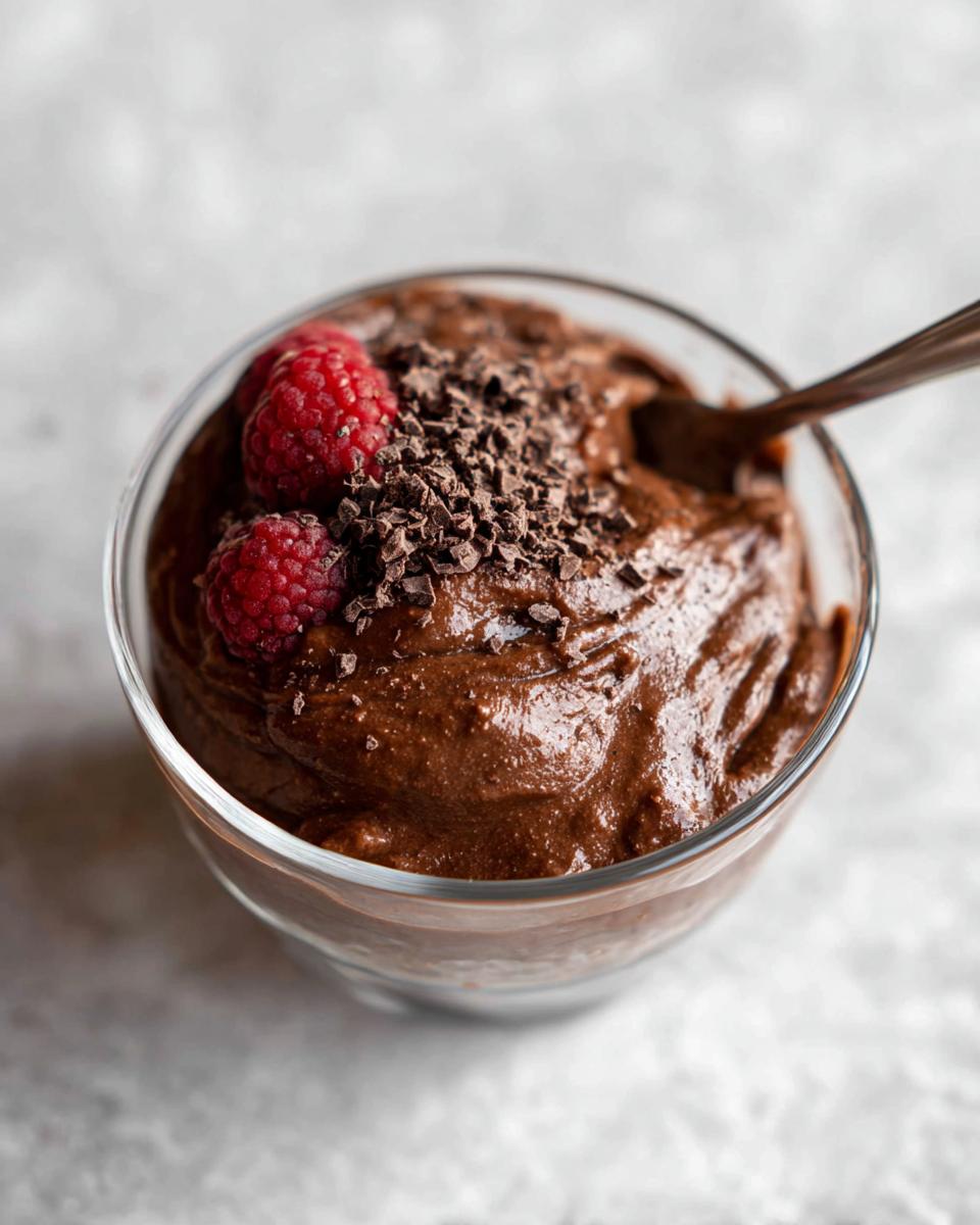 A close-up of a glass bowl filled with rich High Protein Chocolate Pudding, topped with fresh raspberries and chocolate shavings.
