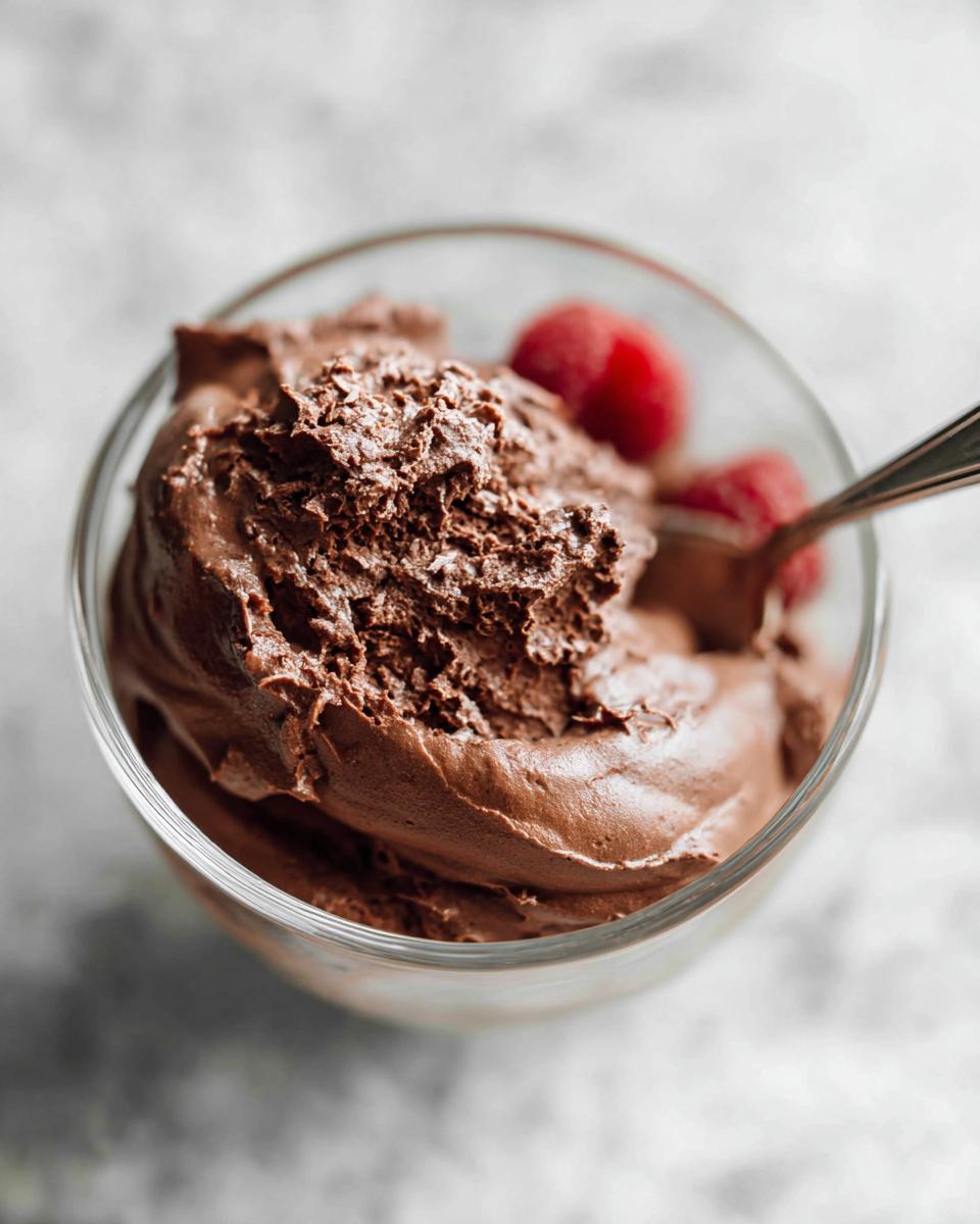 A close-up of a glass bowl filled with fluffy, rich high protein chocolate pudding, topped with fresh raspberries and a spoon.