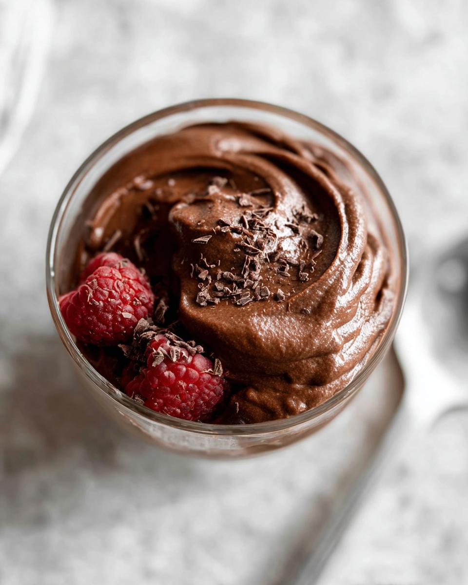 A close-up of creamy High Protein Chocolate Pudding in a glass, topped with chocolate shavings and fresh raspberries.