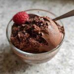 A close-up of a glass bowl filled with decadent High Protein Chocolate Pudding, topped with a fresh raspberry and a sprinkle of sea salt.