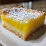 A close-up of a single lemon bar, dusted with powdered sugar, showcasing its bright yellow citrus filling and shortbread crust.