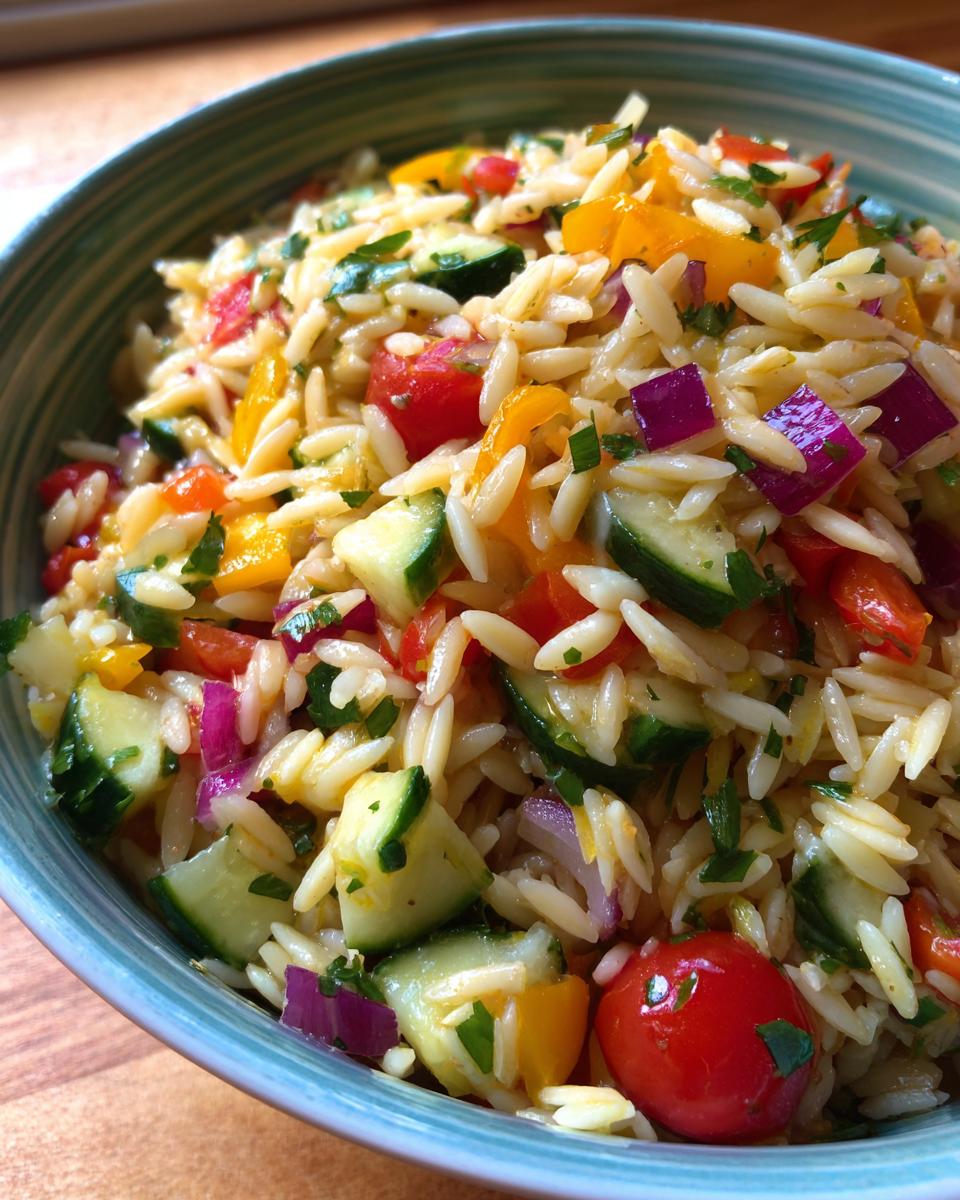A close-up of a bowl of lemony orzo salad with fresh veggies like cucumber, tomatoes, bell peppers, and red onion.