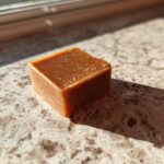 A single square of rich, brown peanut butter fudge sits on a speckled countertop, catching the sunlight.