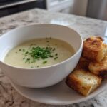 A bowl of creamy potato leek soup topped with chives, served with golden-brown croutons.