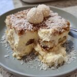 A slice of Raffaello Tiramisu schnell ohne Backen dessert on a plate, topped with coconut flakes and Raffaello candies.