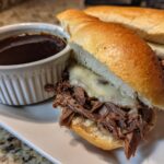 Two slow cooker French dip sandwiches with melted cheese and a side of au jus for dipping.