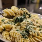 A forkful of creamy spinach artichoke pasta, showcasing fusilli noodles and wilted spinach.