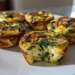 A plate of golden-brown spinach feta egg muffins, with one muffin broken open to show the fluffy interior and spinach filling.